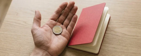 Main tenant une pièce de monnaie à côté d'un livret d'épargne ouvert sur une table en bois clair.