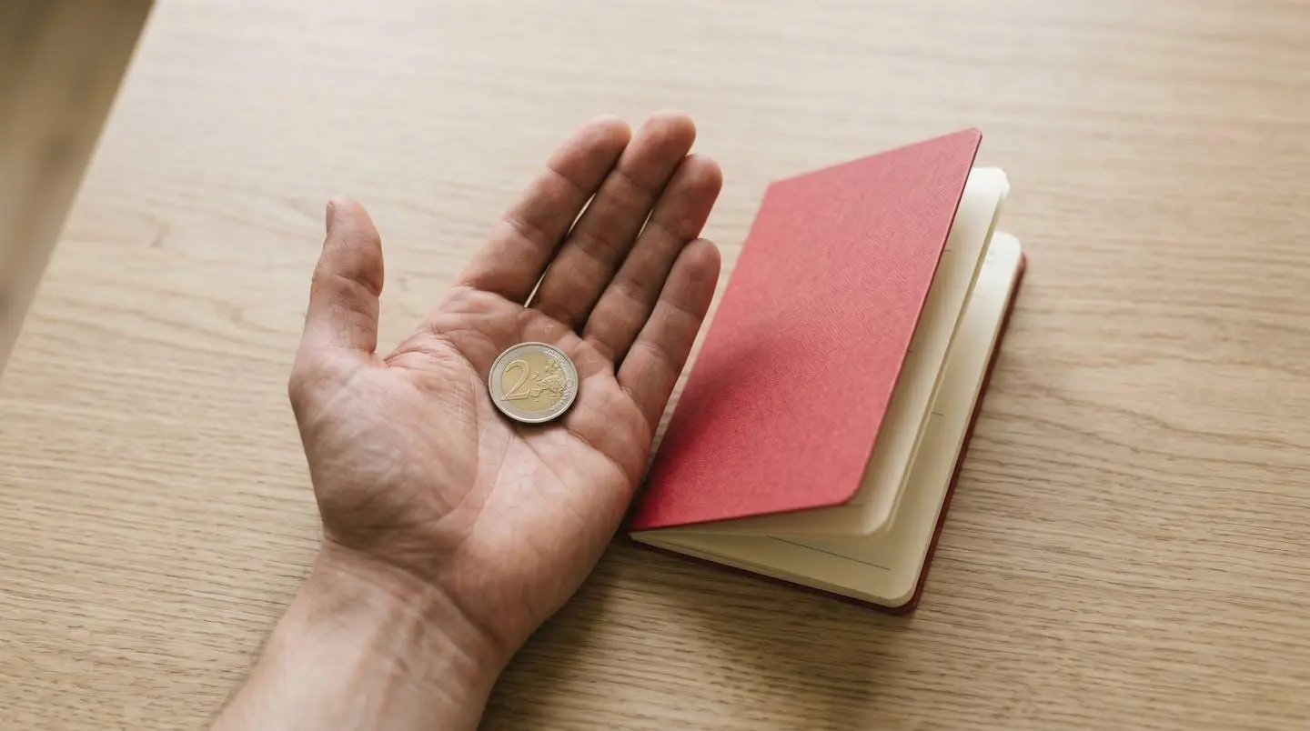 Main tenant une pièce de monnaie à côté d'un livret d'épargne ouvert sur une table en bois clair.